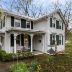 Historic white Victorian farmhouse with dark green shutters and covered front porch at 21 Jay Street in Geneva, NY, featuring decorative gingerbread trim, twin gables, and mature trees in a spring setting.