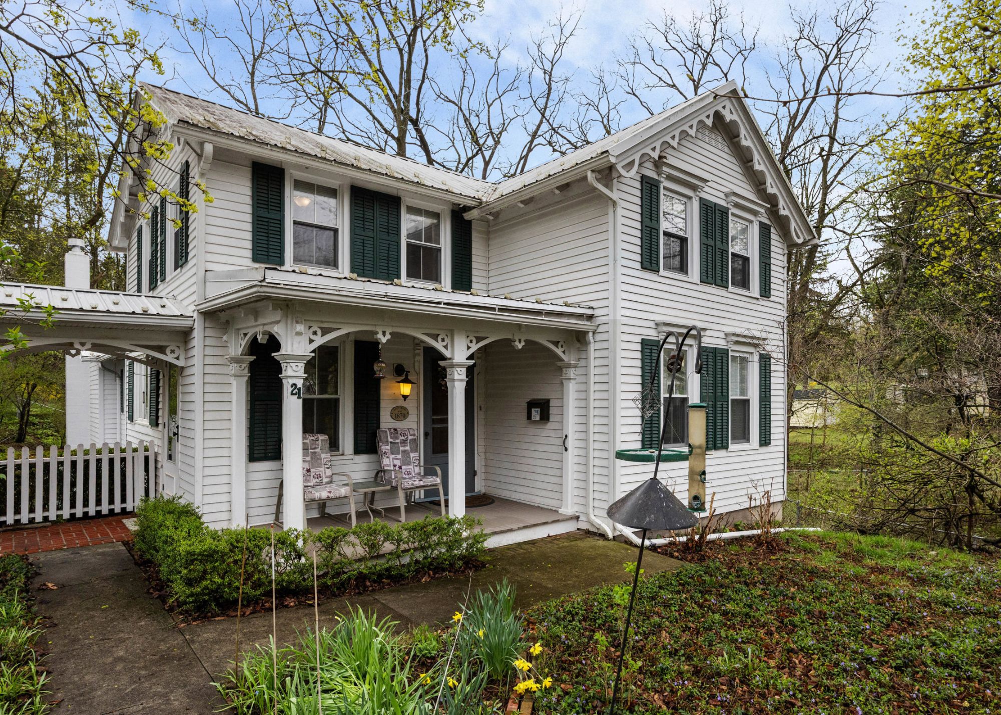 Historic white Victorian farmhouse with dark green shutters and covered front porch at 21 Jay Street in Geneva, NY, featuring decorative gingerbread trim, twin gables, and mature trees in a spring setting.