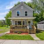 Front exterior of 224 Liberty Street, Oswego NY — two-story gray vinyl-sided home with covered front porch, new wood railings and steps, attic gable vent, and fresh landscaping with mulched bed