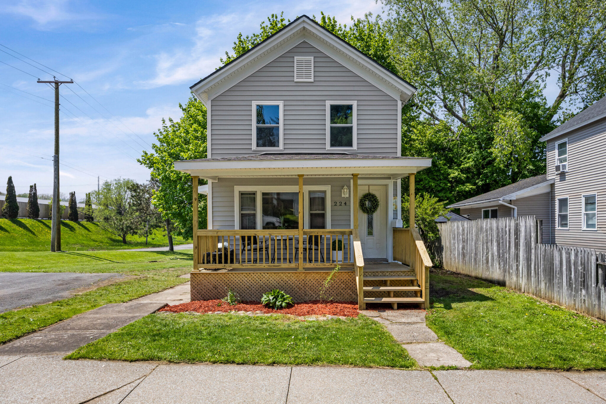 Front exterior of 224 Liberty Street, Oswego NY — two-story gray vinyl-sided home with covered front porch, new wood railings and steps, attic gable vent, and fresh landscaping with mulched bed
